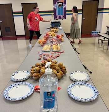 A long table with food, plates, and a hand sanitizer bottle in our school cafeteria. Two women having a conversation stand at the end of the table..