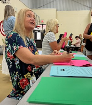 A person seated at a table in a school gymnasium or multipurpose room signs papers placed on bright pink and blue sheets. The person wears a dark floral-patterned top and an ID badge. Another person sits beside them at the same table, also working with papers. Green table coverings, colorful forms, chairs, and several people are visible in the background. A basketball goal and gym walls can be seen behind the registration-style setup.