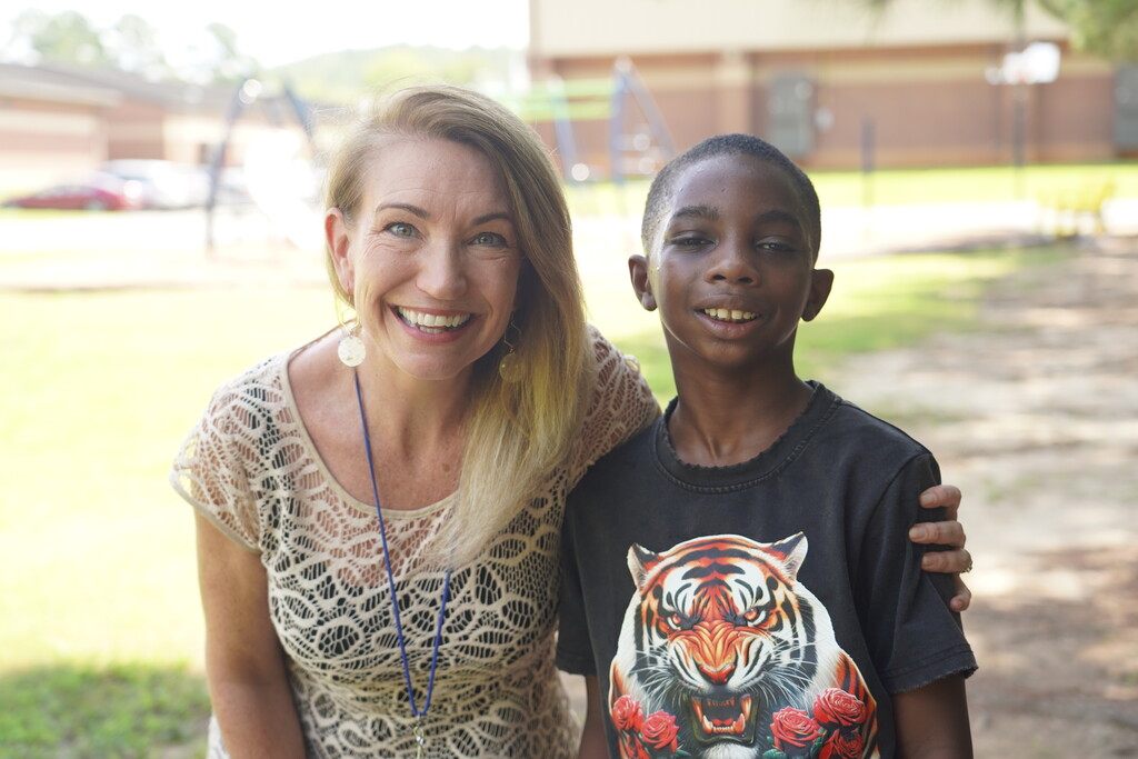 A woman and a boy stand outside, smiling, with a playground and a building behind them.