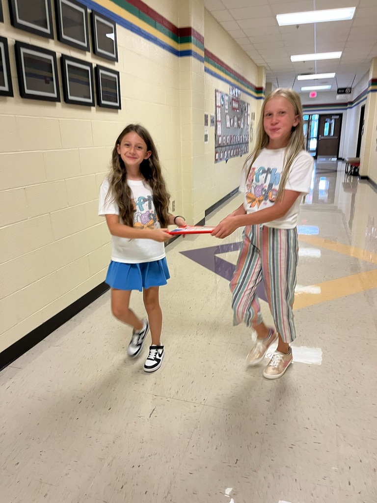 Two smiling students share the task of carrying one composition book in a hallway. Artwork frames, hallway displays, and doors are in the background.