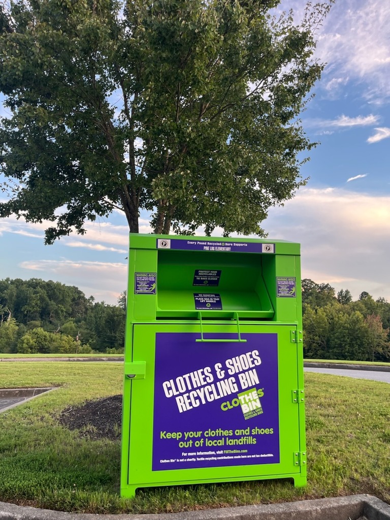 A bright green and purple clothing and shoe recycling bin stands on a grassy area near a parking lot at Pine Log Elementary School. The bin is positioned beneath a large leafy tree and features a large front panel labeled "Clothes & Shoes Recycling Bin." Additional text encourages recycling clothing and shoes to keep them out of local landfills. Trees, grass, and a partly cloudy sky are visible in the background.
