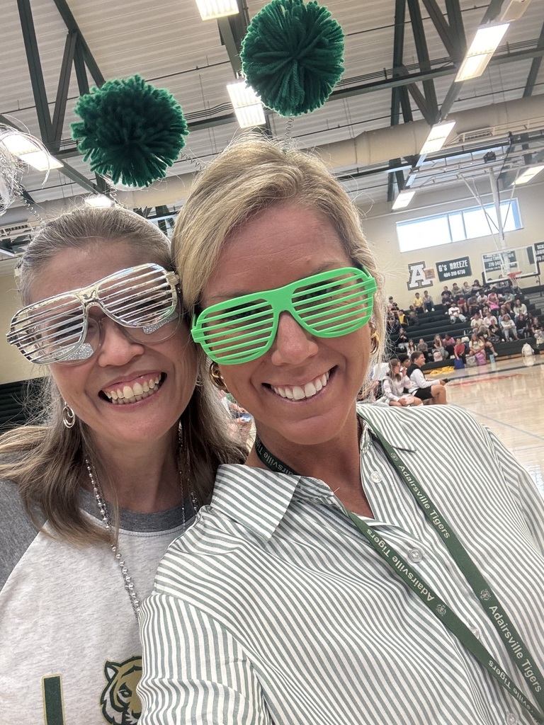 Two women in green sunglasses stand together in a gym, smiling and wearing lanyards.