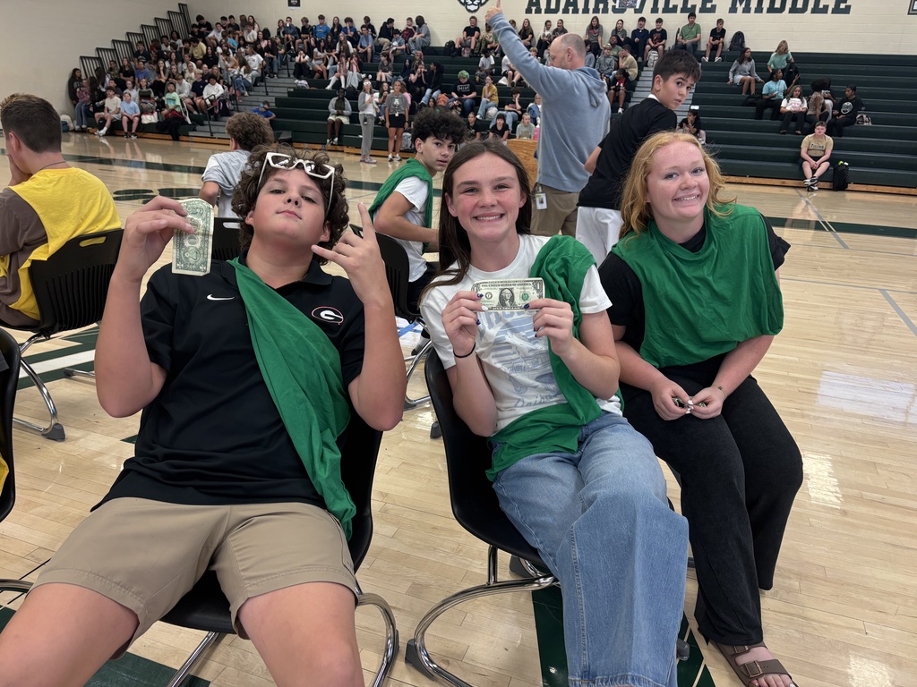 Three students wearing green scarves sit on chairs in a gym with a crowd in the background.
