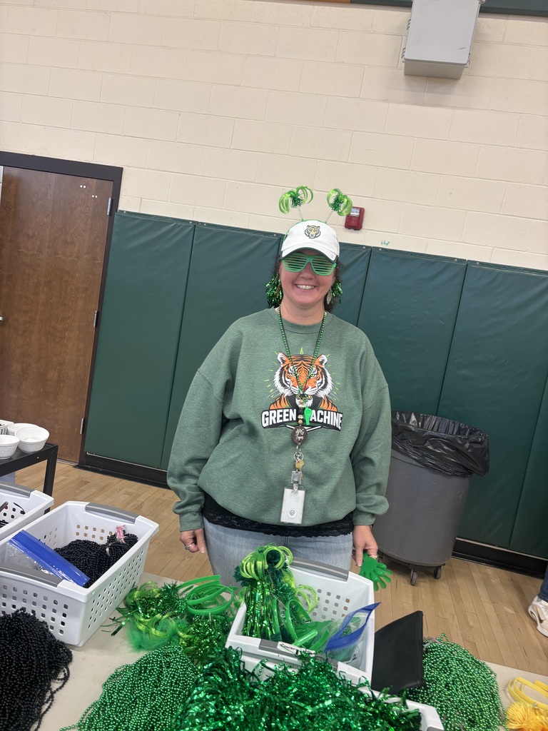 A person in green clothing and a hat stands before a table with baskets and green items.