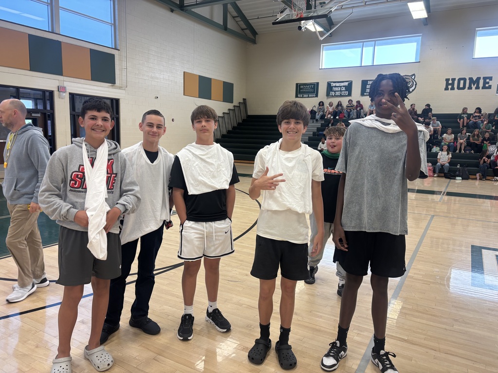 Five boys stand together on a basketball court. Two hold towels around their necks. Behind them, spectators are seated.