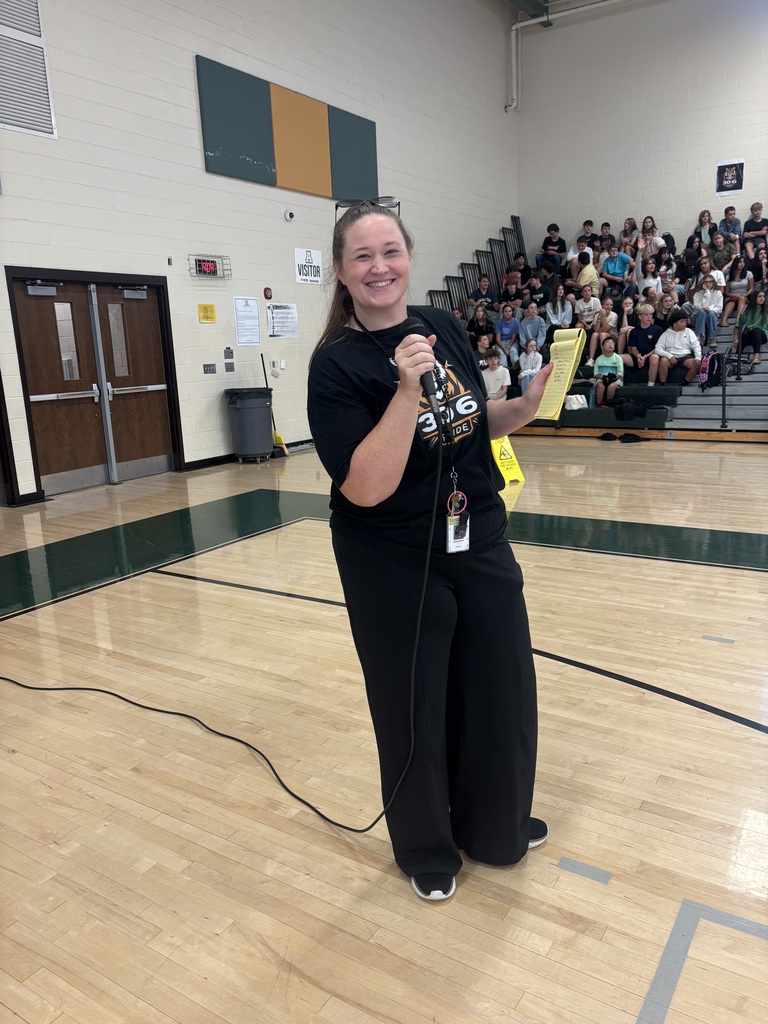 Woman in black outfit holding a microphone stands on a basketball court, with a crowd seated on bleachers.