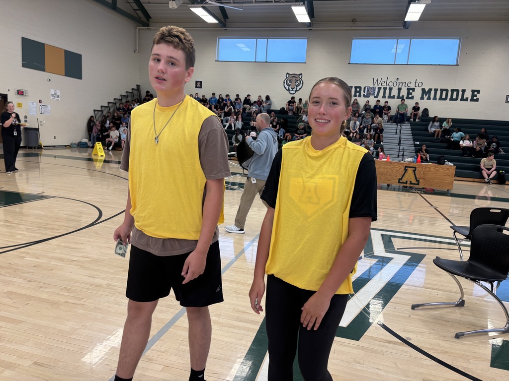 Two individuals, one in yellow and one in blue, stand on a basketball court. Behind them, an audience is seated on bleachers.