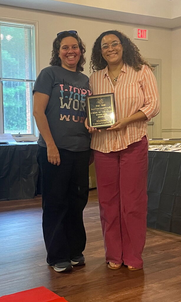 Two women stand in a room, one holding an award plaque, smiling. Behind them, a table with a black cloth, a window with blinds, and an exit sign.