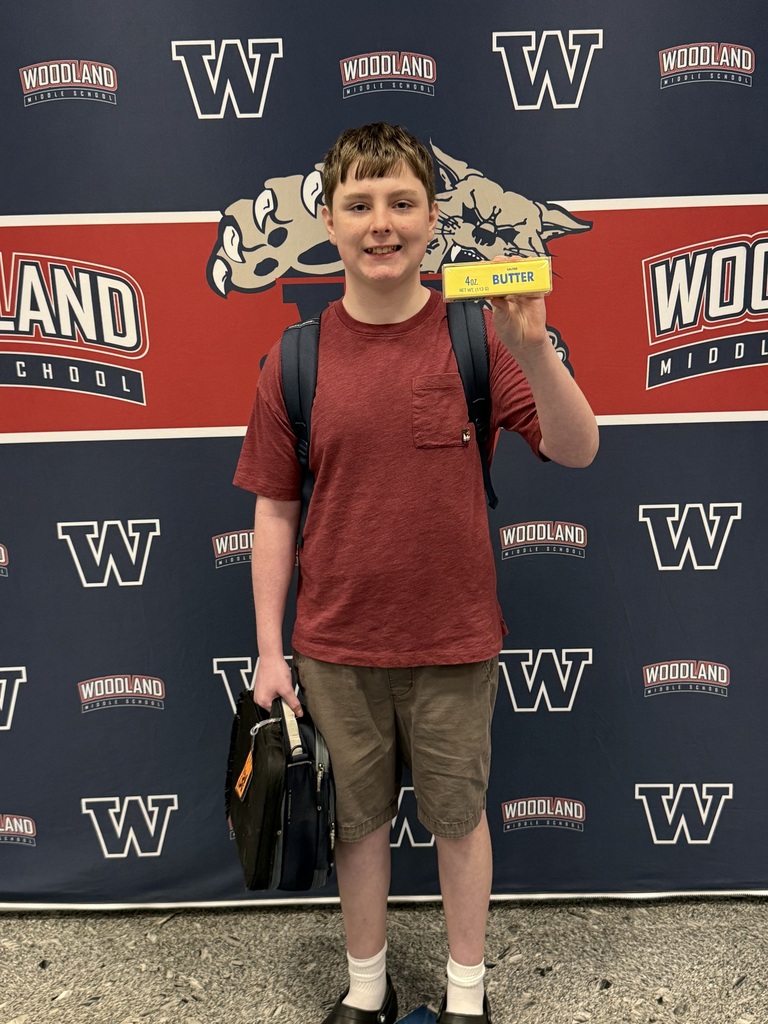 A boy in a red shirt holds a yellow badge, smiling in front of a Woodland school backdrop.