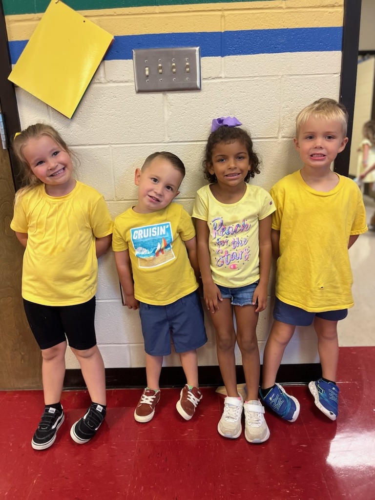 Four smiling children in yellow shirts and denim shorts stand together against the cafeteria wall that has a yellow paper attached to the wall above the children's heads.