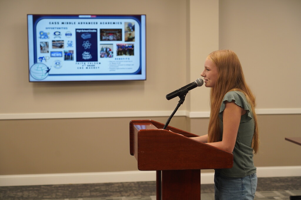Woman with long red hair speaks into a microphone at a podium. Behind her is a large screen with text.