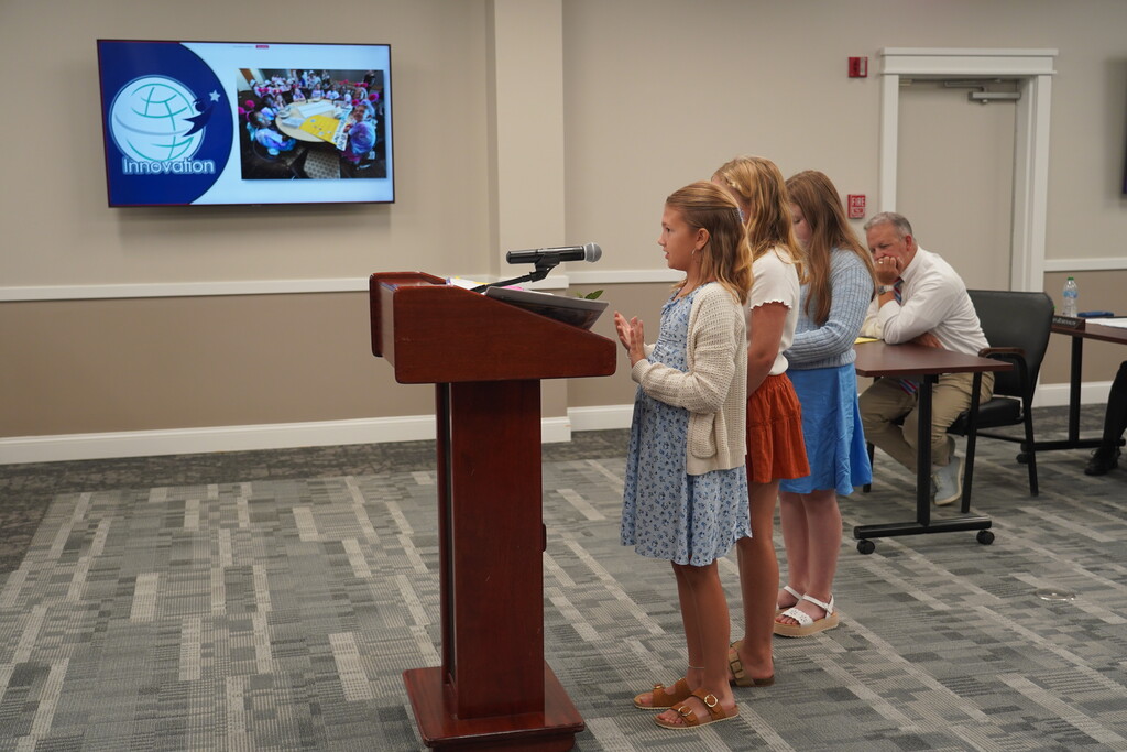 Three girls stand by a podium with a microphone. A man sits at a desk behind them. A monitor displays images.