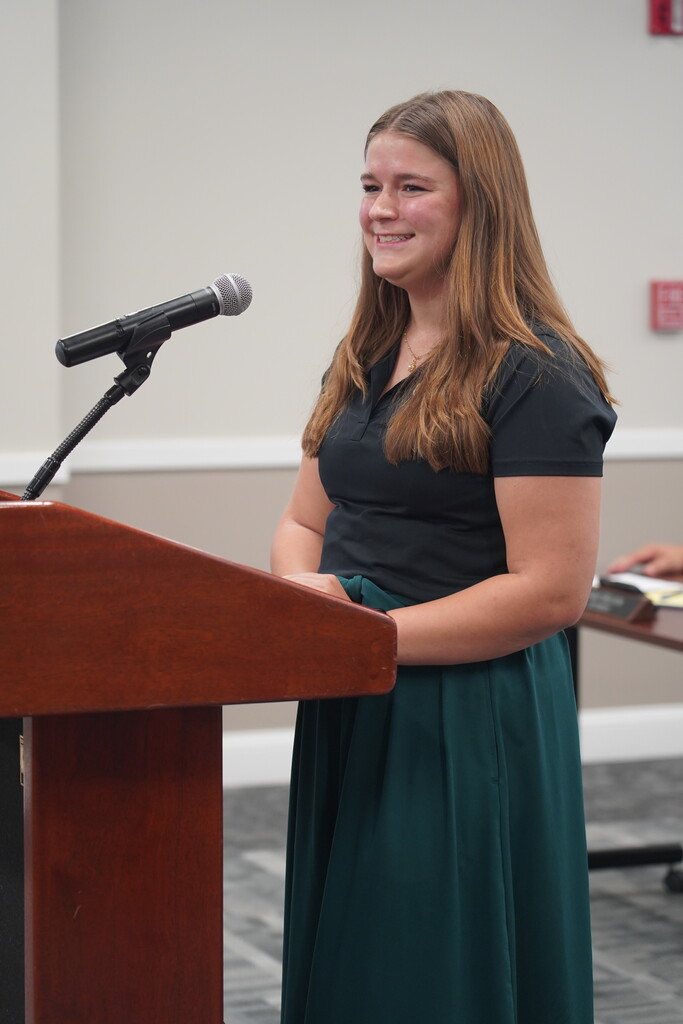 Woman standing at a podium with a microphone in a conference room. She wears a black top and a green skirt.