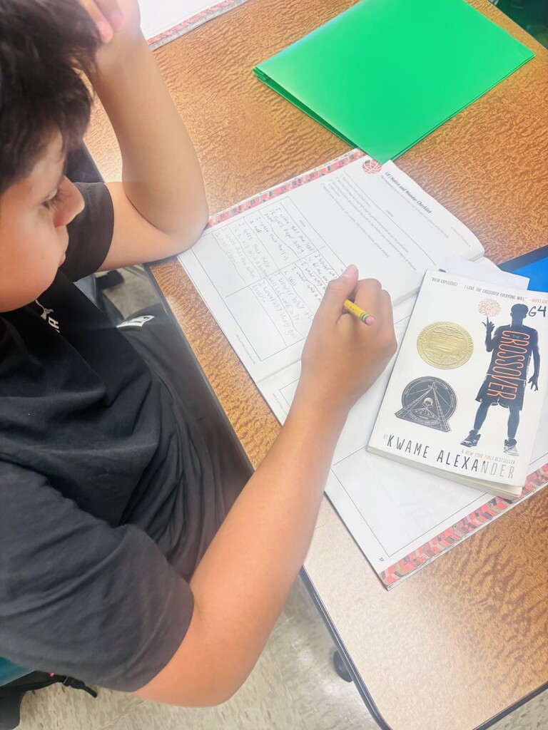 Child seated at a desk, holding a pencil and writing on a book titled "Evaune Alexander." Papers and folders nearby.