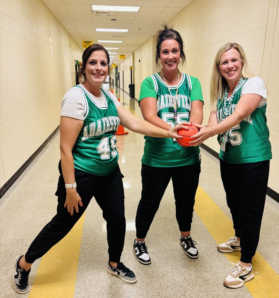 Three women wearing green jerseys with numbers 48, 55, and 85 stand in a hallway, holding a basketball.