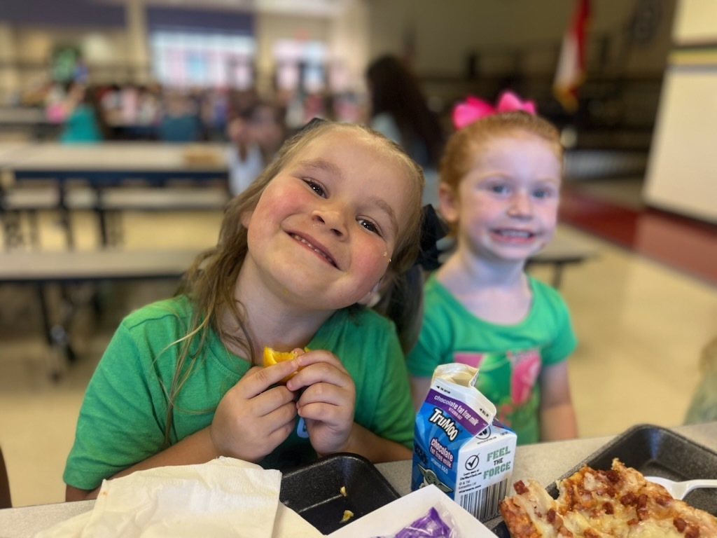 Two Pre-K students in green shirts sit together at a cafeteria table during Green Day, with school lunch trays and classmates in the background.