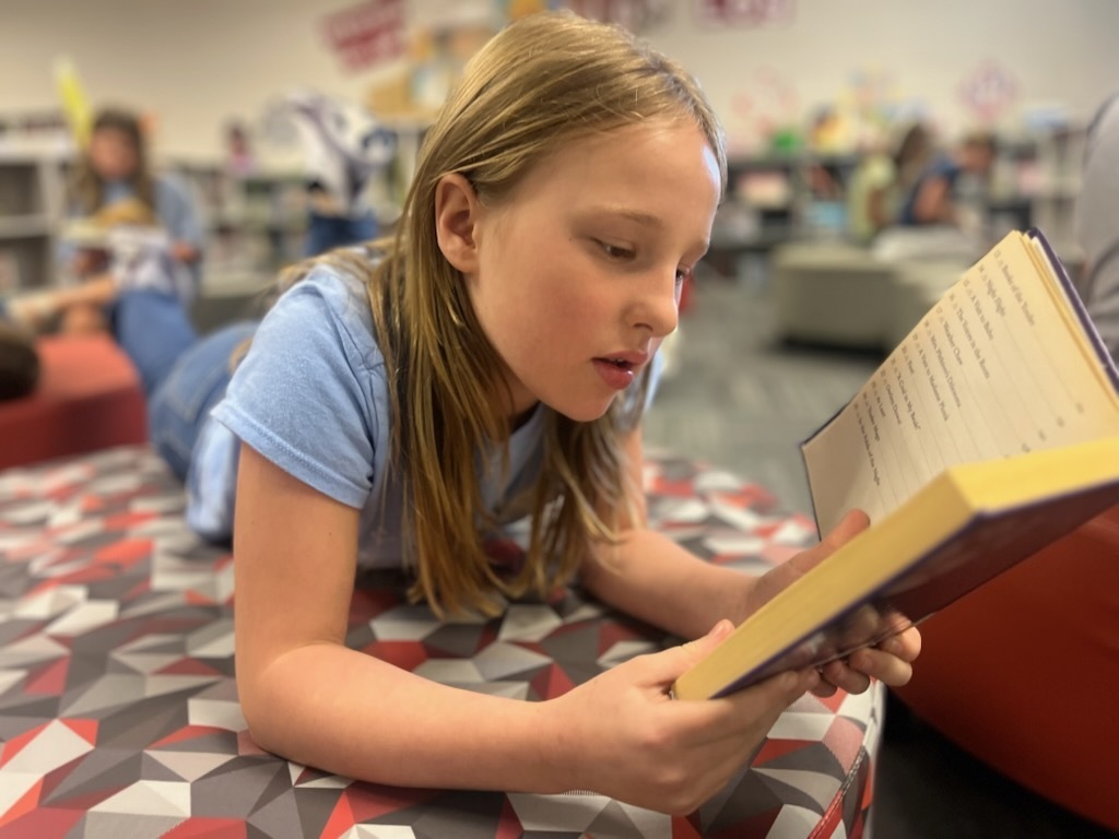 Student engaged in independent reading while relaxing on a cushioned seating area in the library, surrounded by a learning-focused environment.