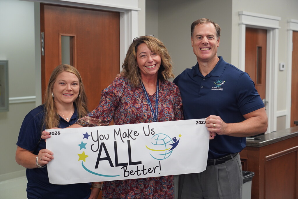 Three people standing and holding a banner that says, "You Make Us All Better!" in a room.