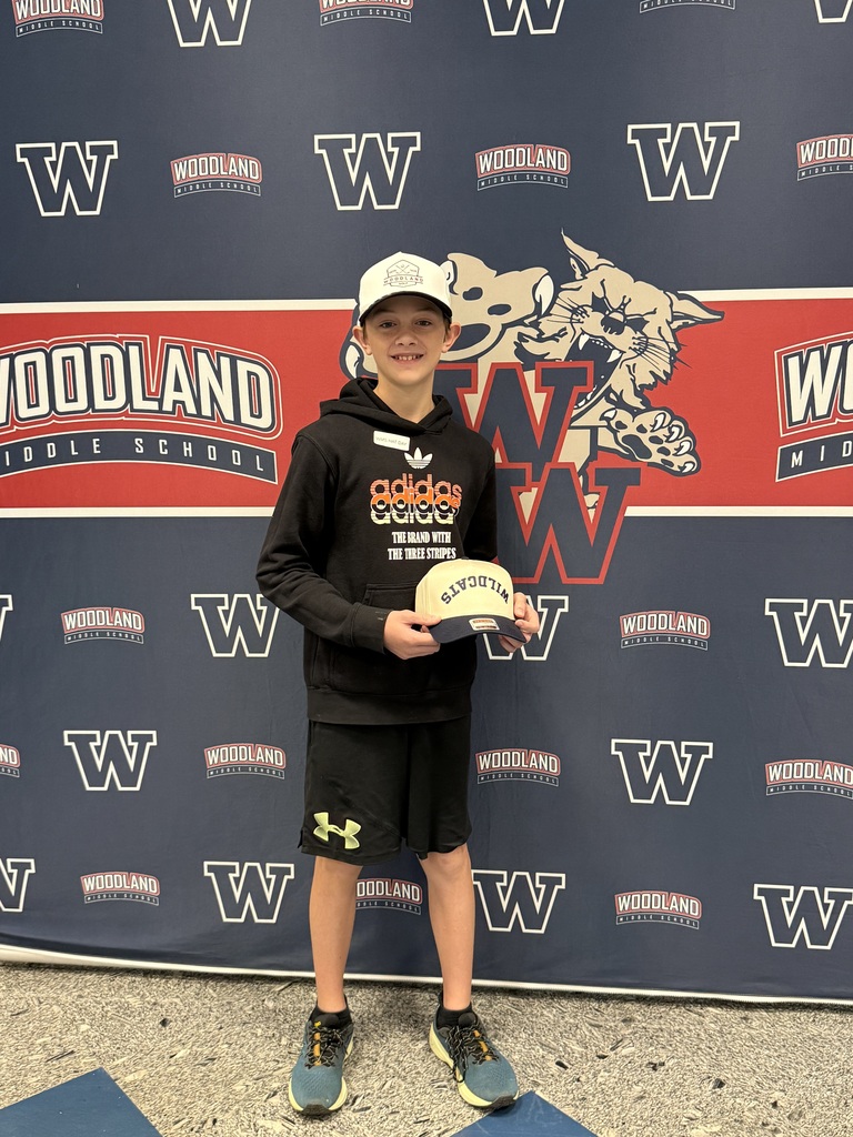 A child in a black hoodie and hat poses with a trophy in front of a Woodland school banner.