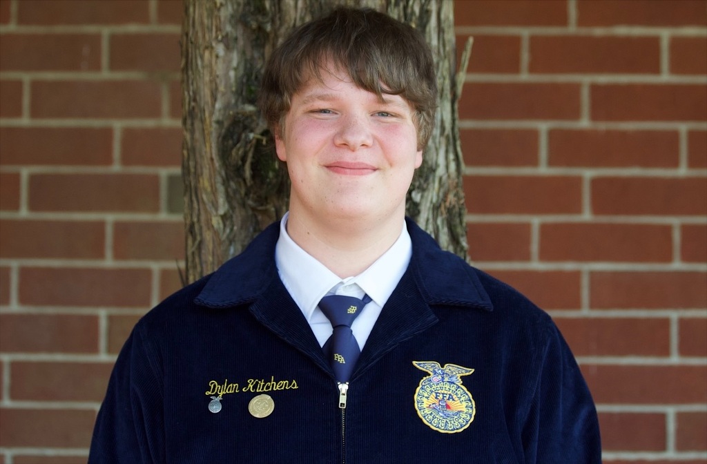 A young man stands smiling in front of a brick wall and a tree, wearing a navy blue jacket and tie.