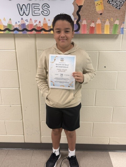A young boy holds a certificate with both hands in a classroom. He wears a tan hoodie and black shorts. Behind him, a wall displays colorful pencils and a sign reading "WES".