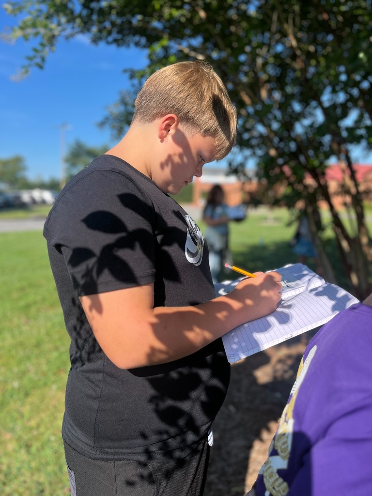 A student in a black shirt writes in a journal outdoors under a tree. A partial view of other students is visible.
