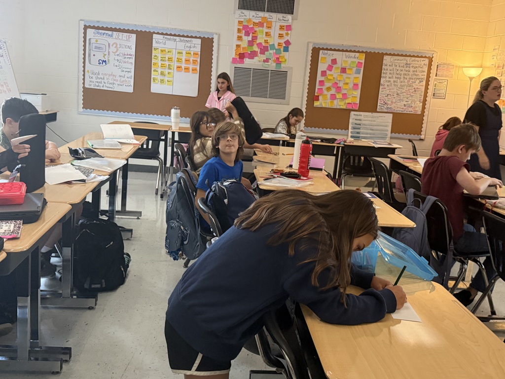 A classroom with students seated at desks, some wearing backpacks. The walls have bulletin boards with various notes and papers.