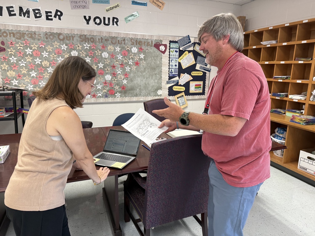 A man and woman stand in a classroom, near a table with a laptop. The man holds paper.