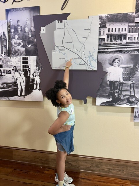 A young girl stands inside, pointing to a map on a wall with historical photographs.