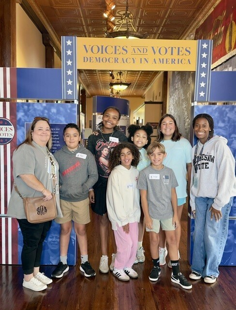 A group of individuals, including adults and children, stand together in a room with a wooden floor.