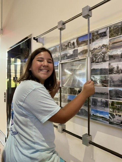 A woman in a white shirt smiles at a wall with photos on a metal frame.