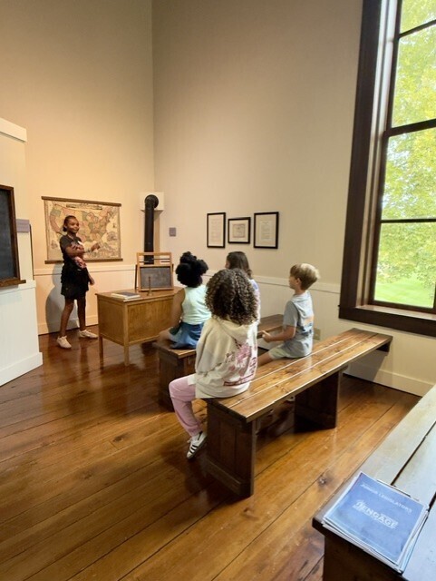 A room with wooden floors, a person standing, children sitting on benches, and a window with trees visible.
