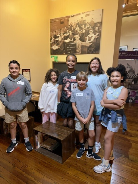 Group of children in a room, some with arms crossed, smiling at camera. A wooden box and desk.