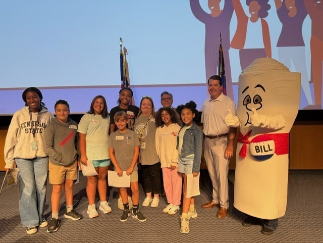 Group of kids and adults stand together for a photo in front of a screen. One person is a mascot.