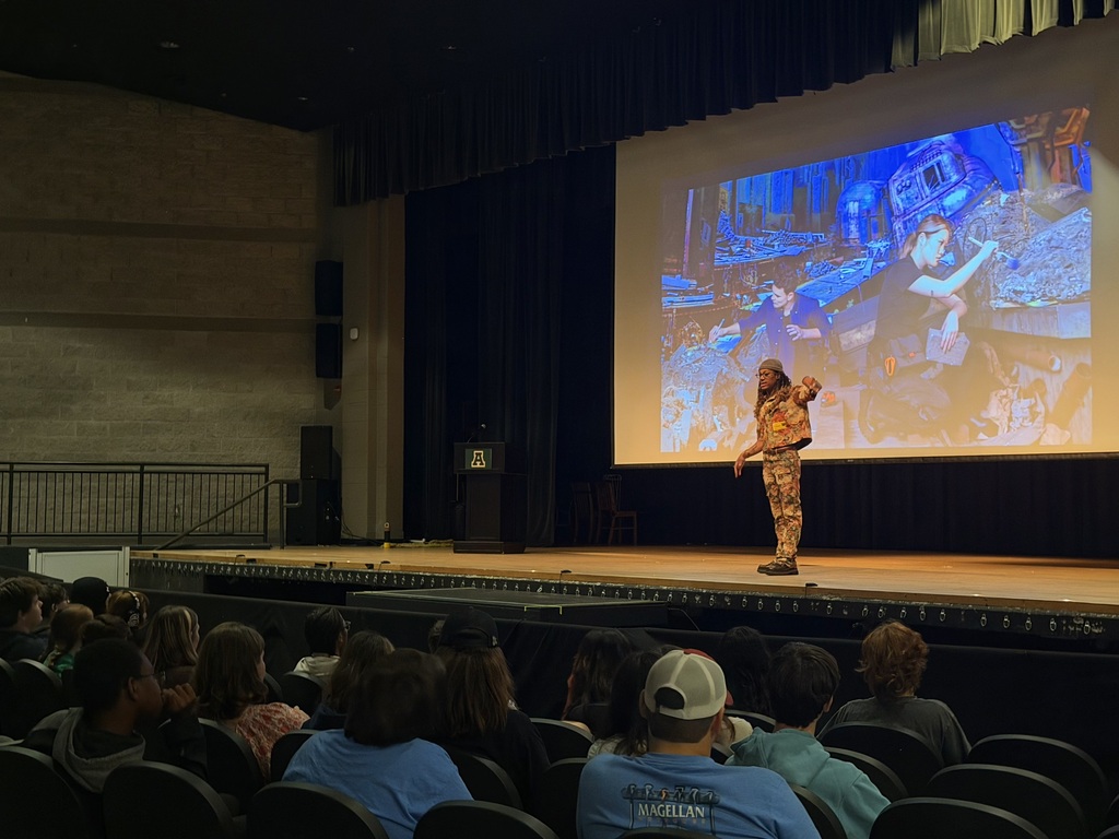 An auditorium with an audience seated, a person in camouflage speaks on stage, a projector screen behind them shows an image.