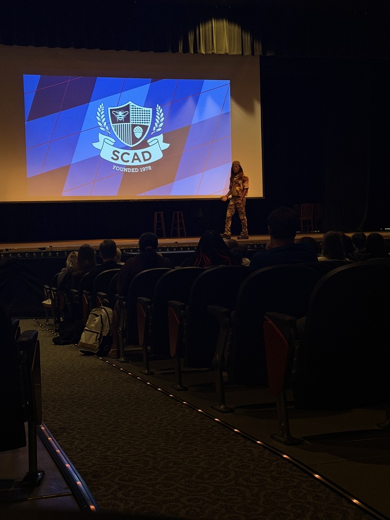 A person on stage in front of an audience. The stage has a large screen behind the speaker.