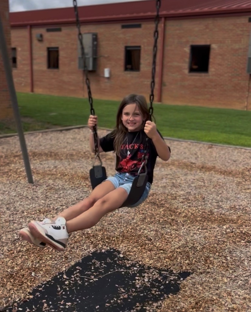A young child sits on a swing with legs pushed out on a playground with a red-roofed building behind her.