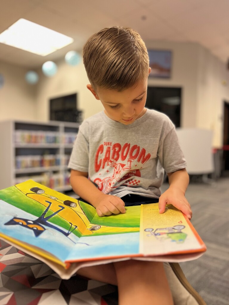 Student seated in the school library, focused on reading an open Pete the Cat picture book with colorful illustrations. Library bookshelves and learning spaces are softly blurred in the background.