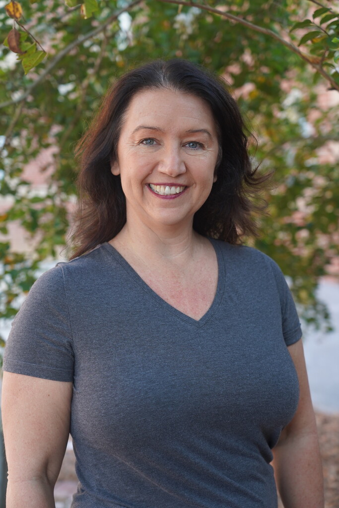 A woman smiles, standing near a tree. She wears a blue V-neck shirt, and her hair falls to her shoulders.