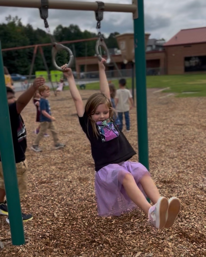 Student hanging from a set of overhead playground rings during recess on a school playground. Other students are visible in the background, with swings and school buildings behind them.