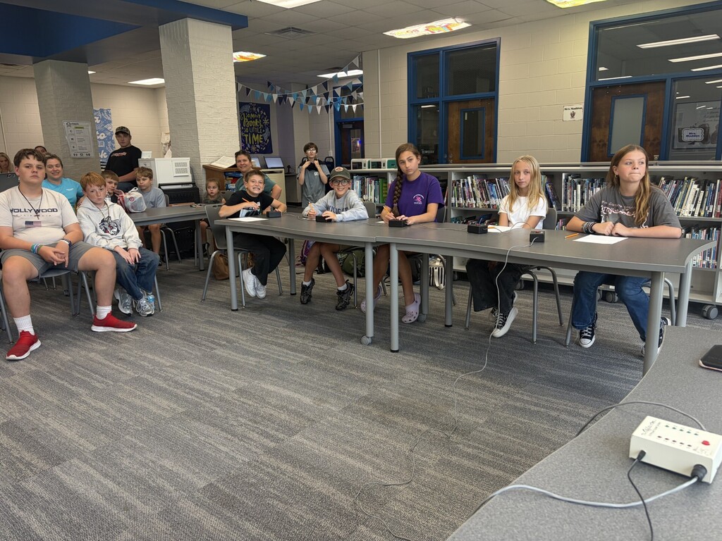 A group of students sitting at tables in a library, some with papers and microphones.