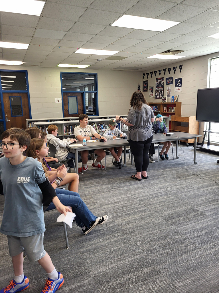 A room with tables and chairs, a person speaking at the front, and several students sitting and standing.