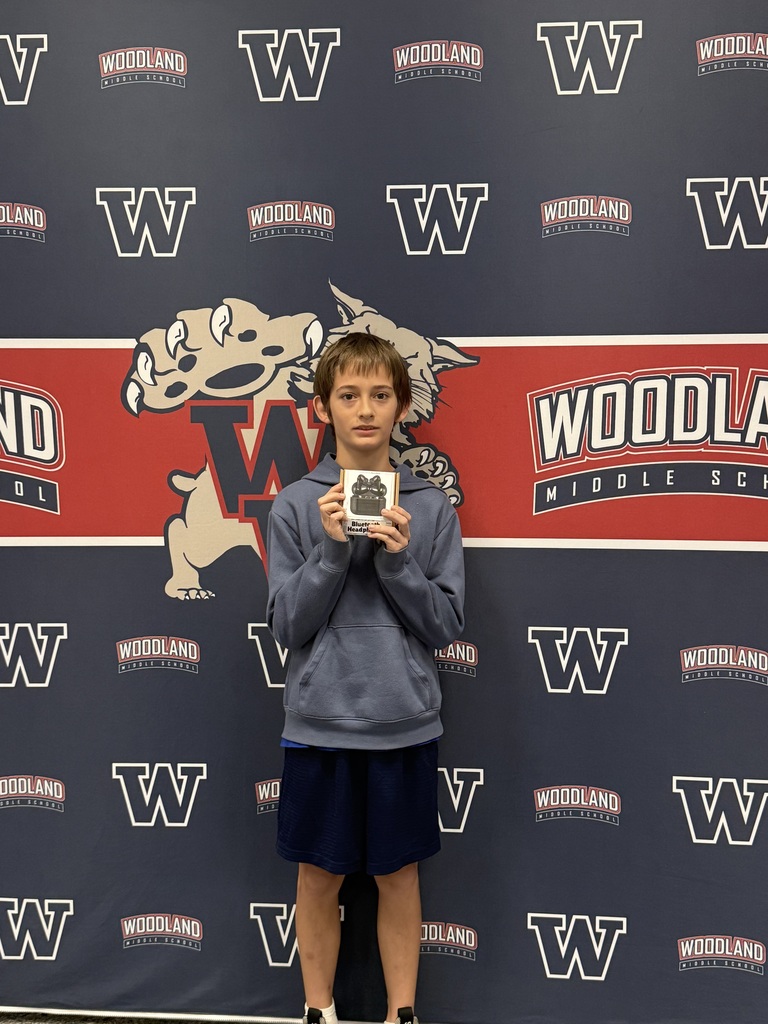 Person in a gray shirt and blue skirt holds a small trophy in front of a wall with Woodland Middle School logos.