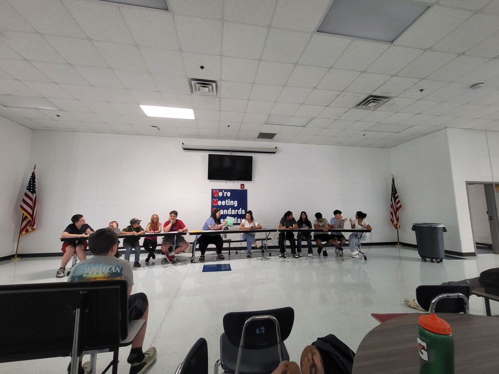 Several students sit at tables with a US flag and another flag in the background.