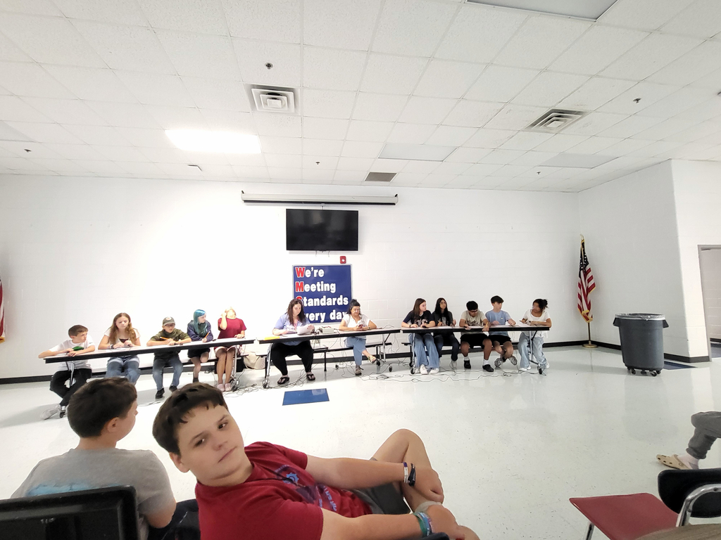 A classroom with tables and chairs. Several students sit at tables, and one student sits on a chair.