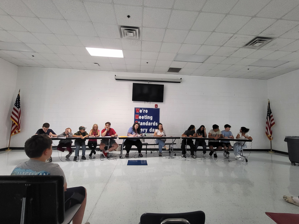 A group of people seated at tables in a room with white walls and flags. A blue banner is displayed behind them.