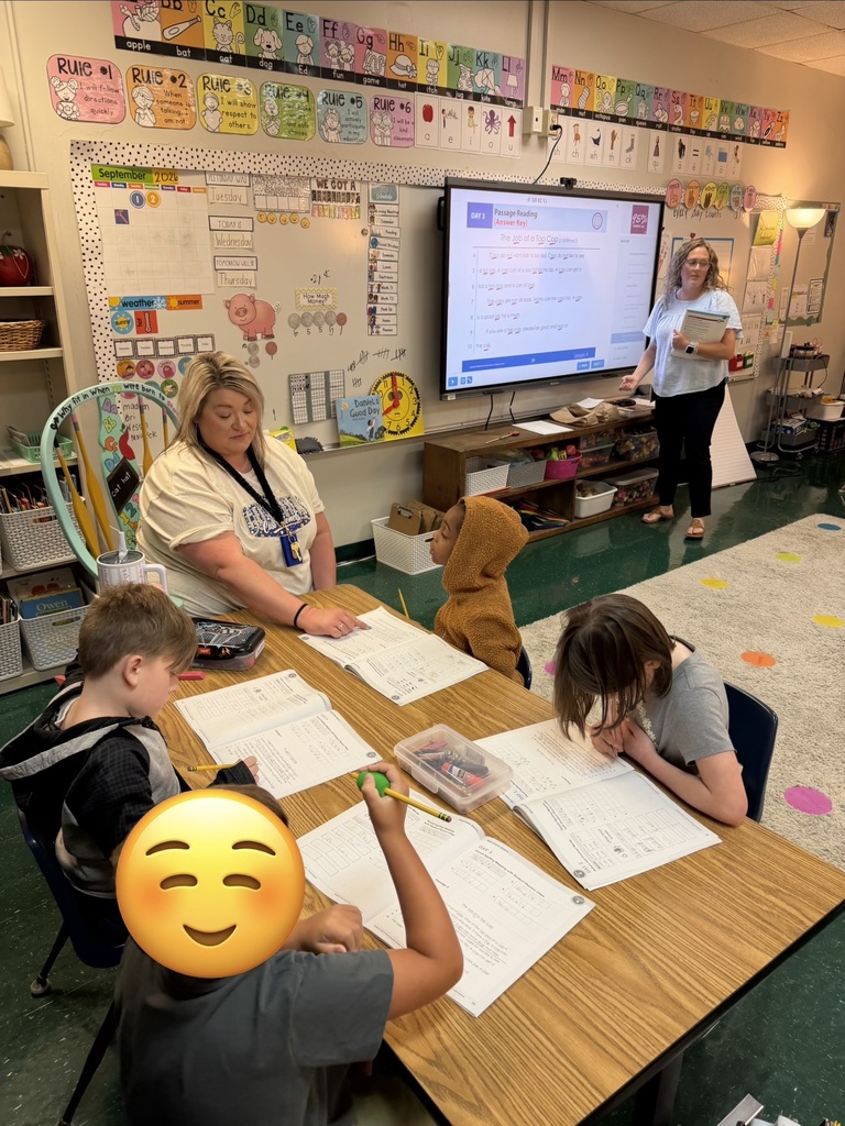 Four children sit at a table in a classroom. Papers, pens, and pencils are on the table.