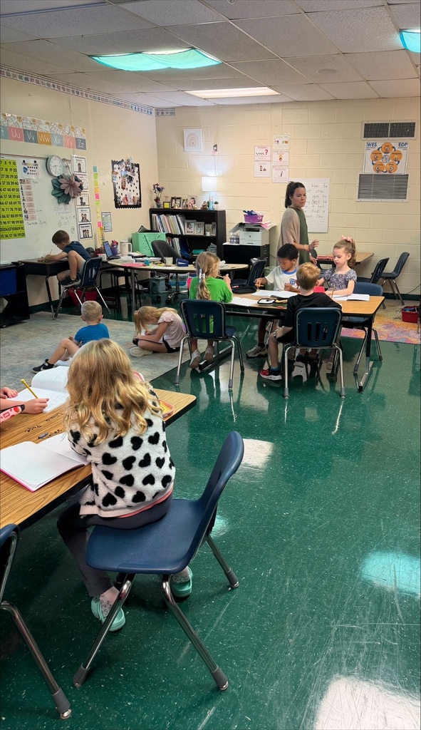 A classroom with green floors and white walls. Students sit at desks and some are standing. A teacher is in the middle.