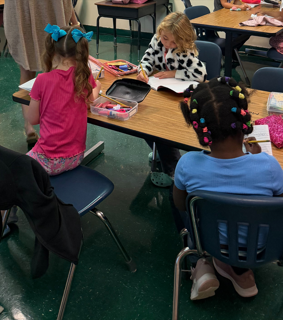 Three young children seated at a table, engaged in activities. One holds a book, and others have art supplies.