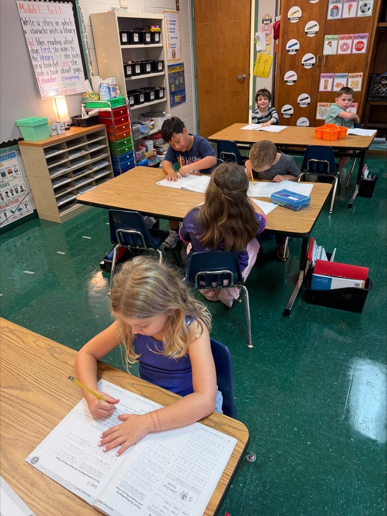 A classroom with green tile floors and wooden desks. Children sit at tables, using pencils to write in notebooks.